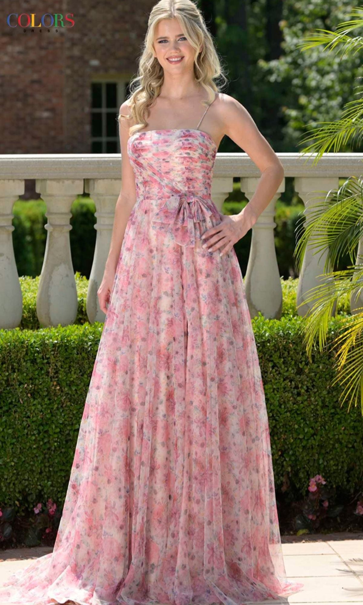 Woman poses outdoors by a stone railing in the Colors Dress 3812—a strappy, long pink prom dress with floral print and bow at waist—surrounded by lush greenery.
